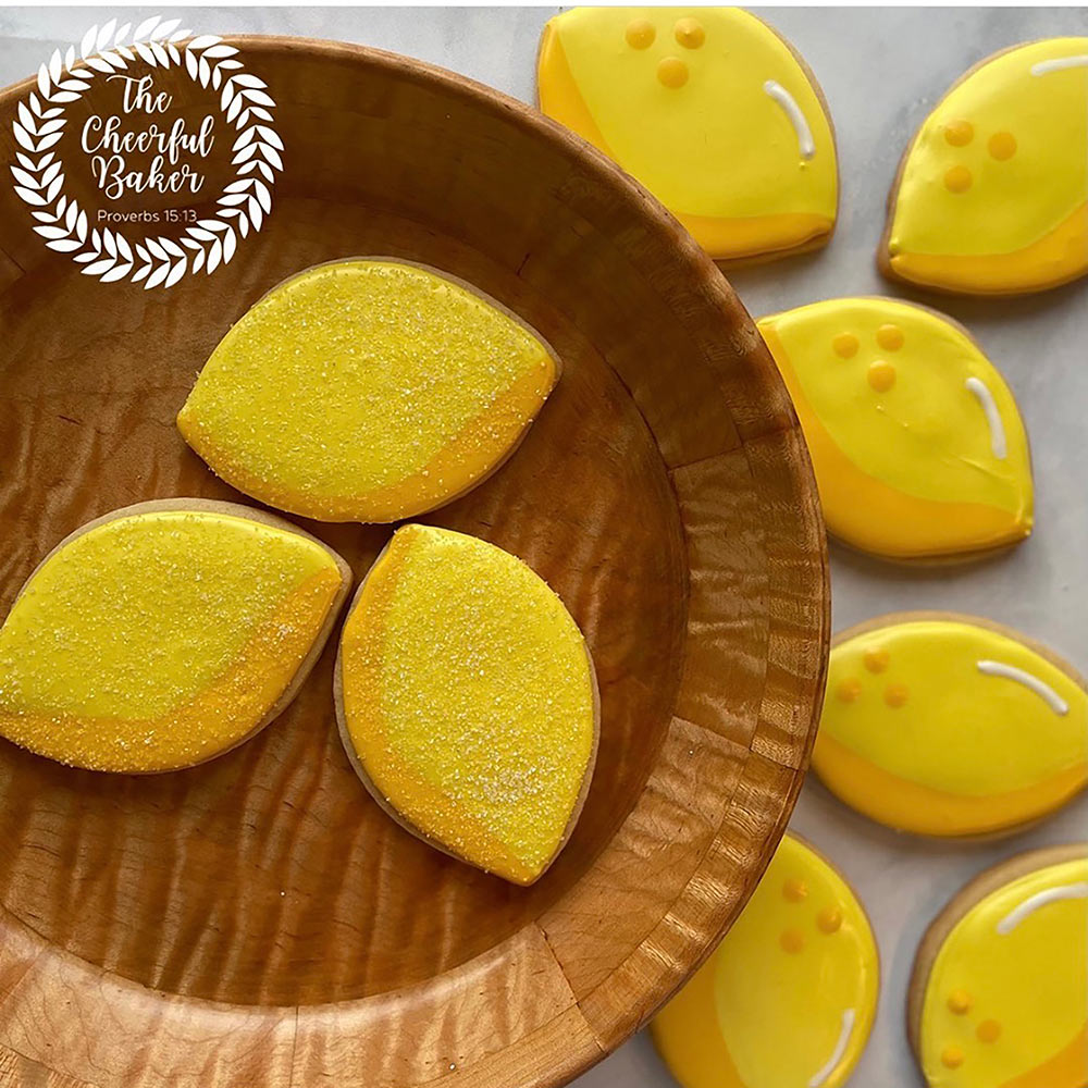 three lemon shaped and decorated cookies on a wooden plate, with a bunch of other lemon shaped cookies beside it