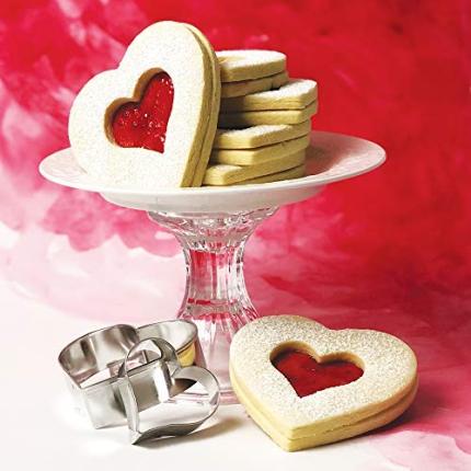 Heart shaped linzer cookies on an elevated plate with red jam, cookie cutters below the plate,  and a red watercolor background