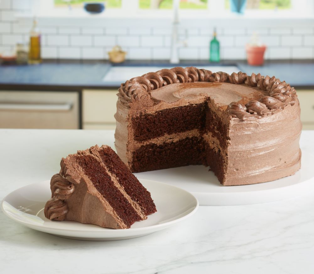 Image of a slice of chocolate cake with chocolate frosting sitting in front of the full chocolate cake.