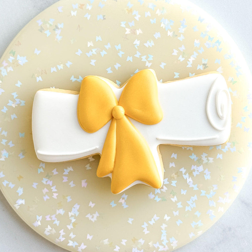 Image of a diploma sugar cookie decorated with white royal icing and a gold bow.