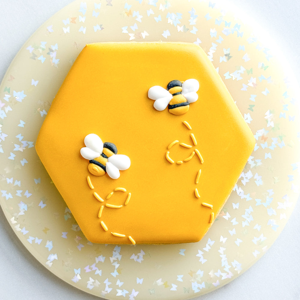 Image of a hexagon shaped cookie decorated with yellow royal icing and bee royal icing transfers.