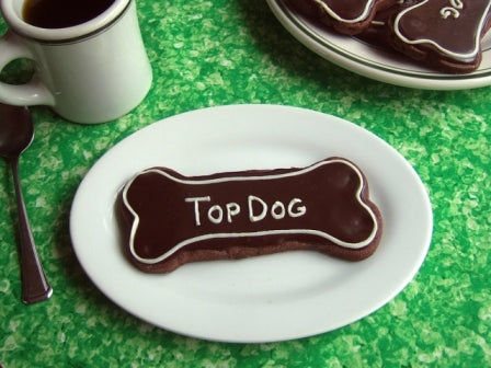 chocolate dog bone cookie that says top dog in royal icing on a white plate and green tablecloth