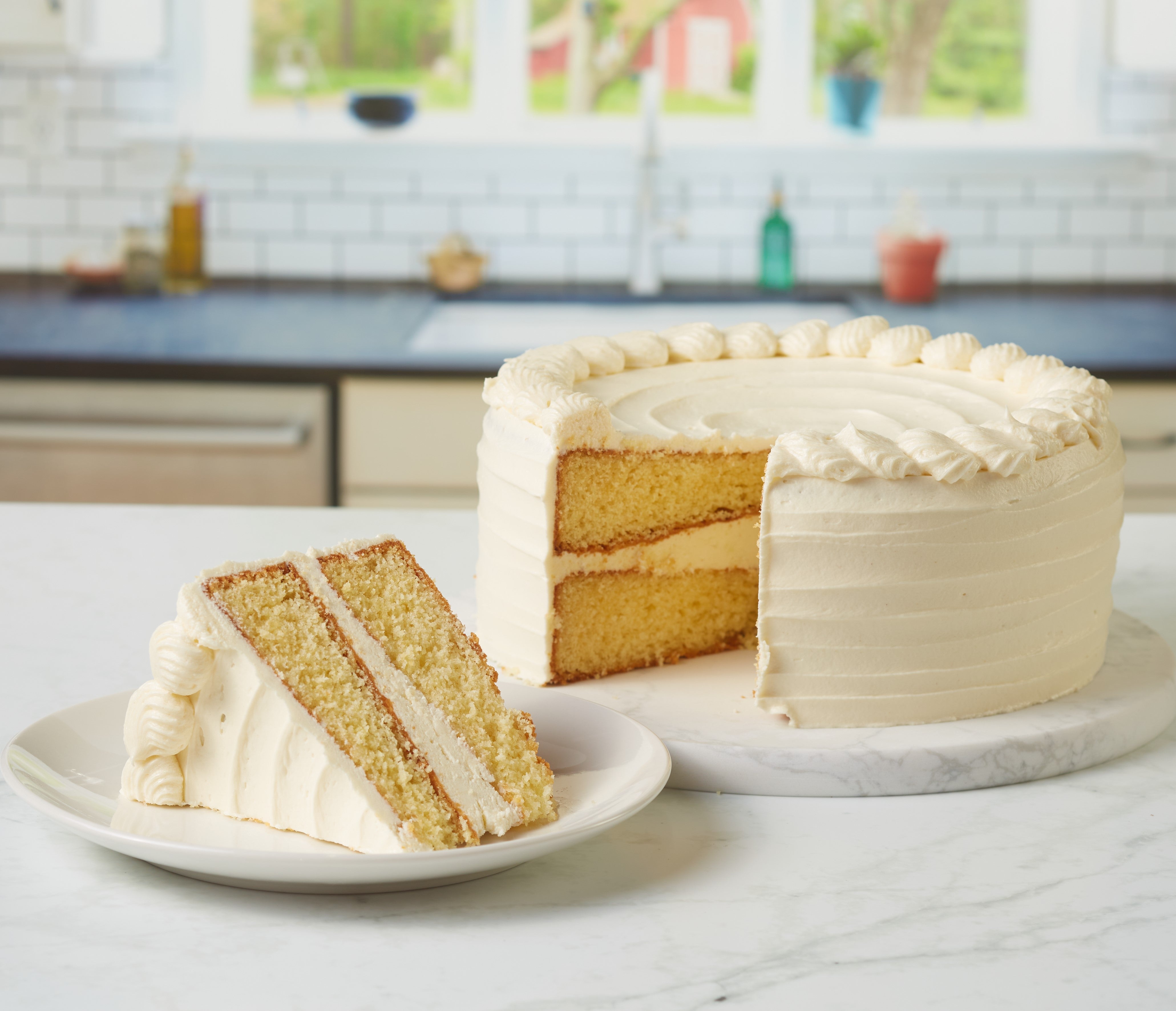 Image of a slice of yellow cake with vanilla frosting, with the full cake in the background.
