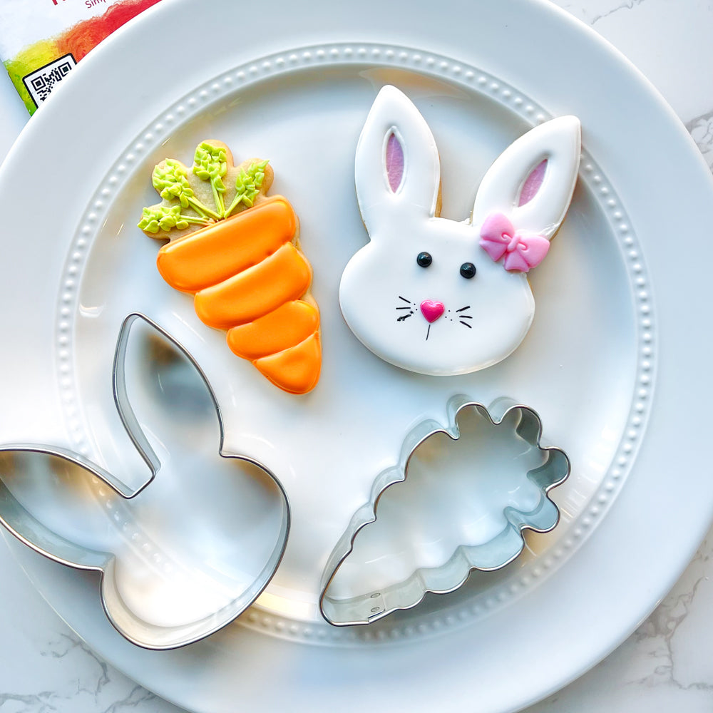 Image of an Easter bunny decorated sugar cookie and carrot sugar cookie alongside cookie cutters on a white plate.