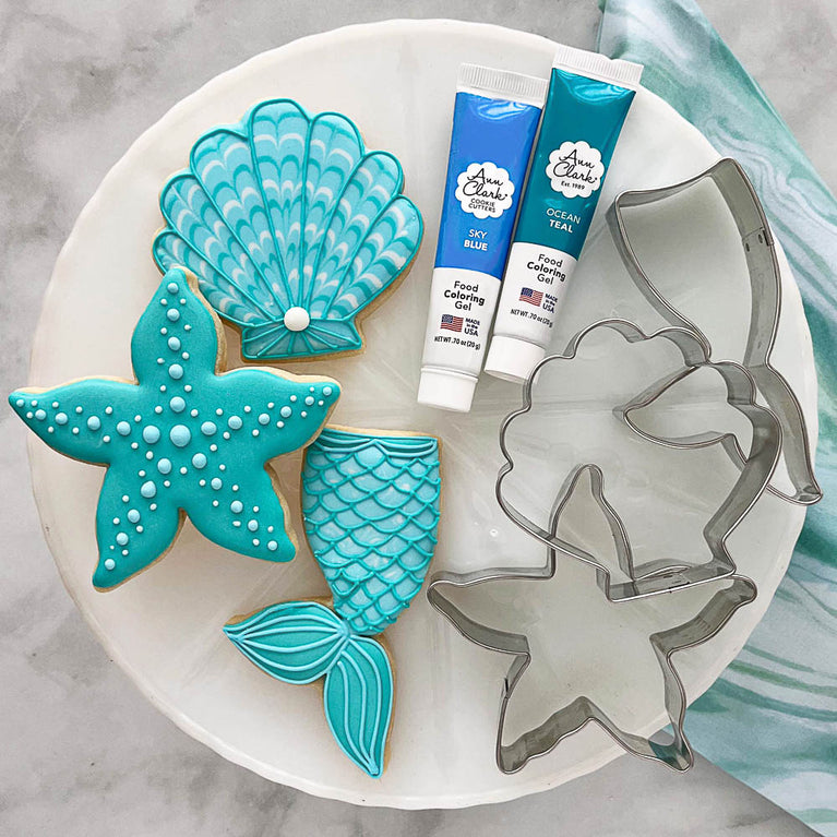Mermaid cookies on a plate with cookie cutters and food coloring gel tubes